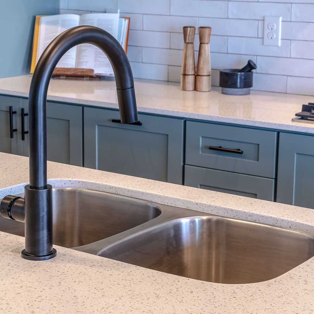 Square frame Double bowl sink and black faucet on kitchen island against cooktop and counter. Cooking appliances and cabinets against white tile backsplash can be seen in the background.